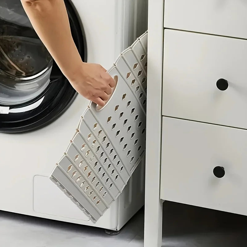A hand is placing a folding laundry basket with handles into a white cabinet drawer.