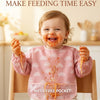 A smiling baby wearing a pink bib with a mess-free pocket, covered in food, sitting in a wooden chair.