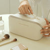 A hand is opening a cream-colored leather travel makeup organizer bag on a white surface, with makeup brushes and a compact p