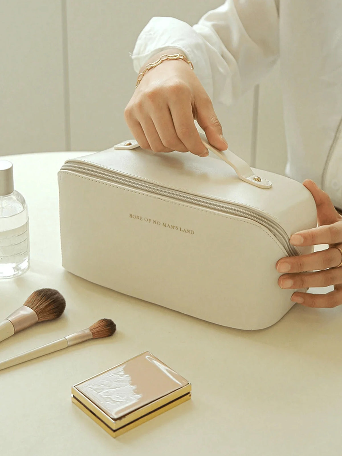 A hand is opening a cream-colored leather travel makeup organizer bag on a white surface, with makeup brushes and a compact p