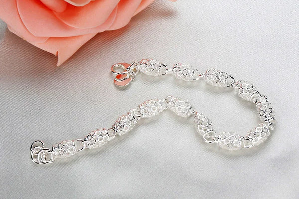 Una delicada pulsera de cadena geométrica de plata esterlina yace sobre una superficie blanca junto a una rosa rosa.