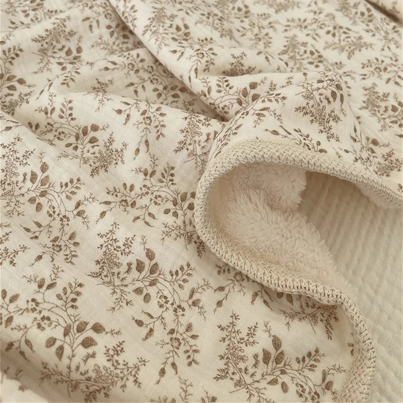 Close-up of a floral muslin baby blanket in beige with brown botanical prints, showing soft texture and folds.