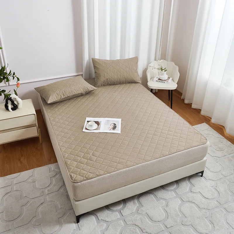 A beige waterproof mattress protector with elastic straps is displayed on a bed, next to a white chair and a light-colored ru