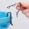 A hand holds black plastic clips designed to secure garbage bags or snack bags onto a trash can.