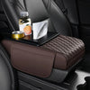 A dark brown quilted car armrest cushion with storage and cup holders is placed on a black car seat.
