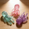 Three soft silicone glitter squishy toys in shades of teal, pink, and purple resting on a wooden surface.