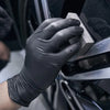 A hand wearing a black nitrile cleaning glove is using a wooden tool to clean a dark surface.