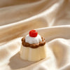 A small white cake with a chocolate ring and a red cherry on top resting on a beige fabric.
