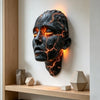 A dark, cracked face sculpture illuminated by warm orange light mounted on a white wall.