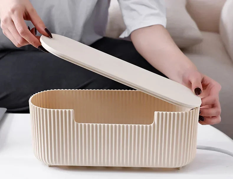 A hand opens a light beige cable management box organizer for power strips on a white surface.