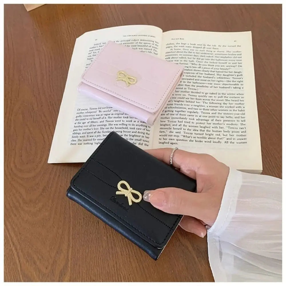 A black tri-fold wallet with a gold bow detail is shown resting on an open book.
