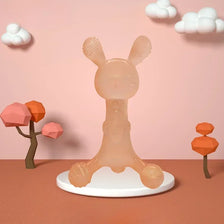 A light peach silicone cartoon rabbit teether standing on a white plate against a pink background with clouds and orange geom