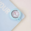 A light blue silicone pocket watch with a silver bezel and a pink-colored brooch pin is shown on a light blue background.
