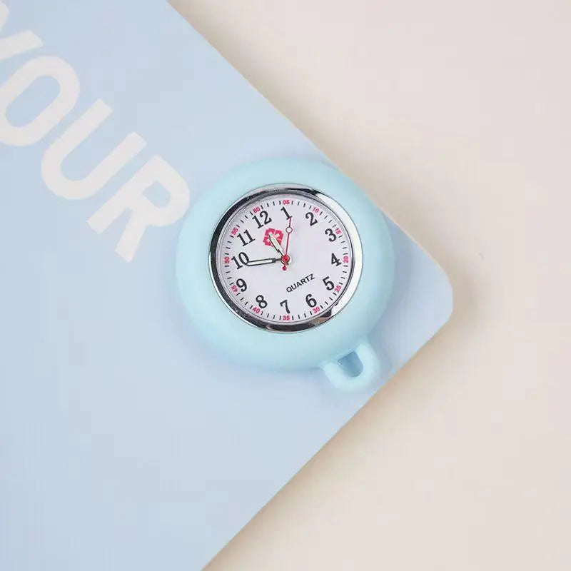 A light blue silicone pocket watch with a silver bezel and a pink-colored brooch pin is shown on a light blue background.