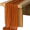 A rust orange cotton gauze table runner draped over a wooden table.