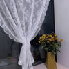 White floral lace sheer curtain panel draped over a glass surface with a vase of yellow flowers.