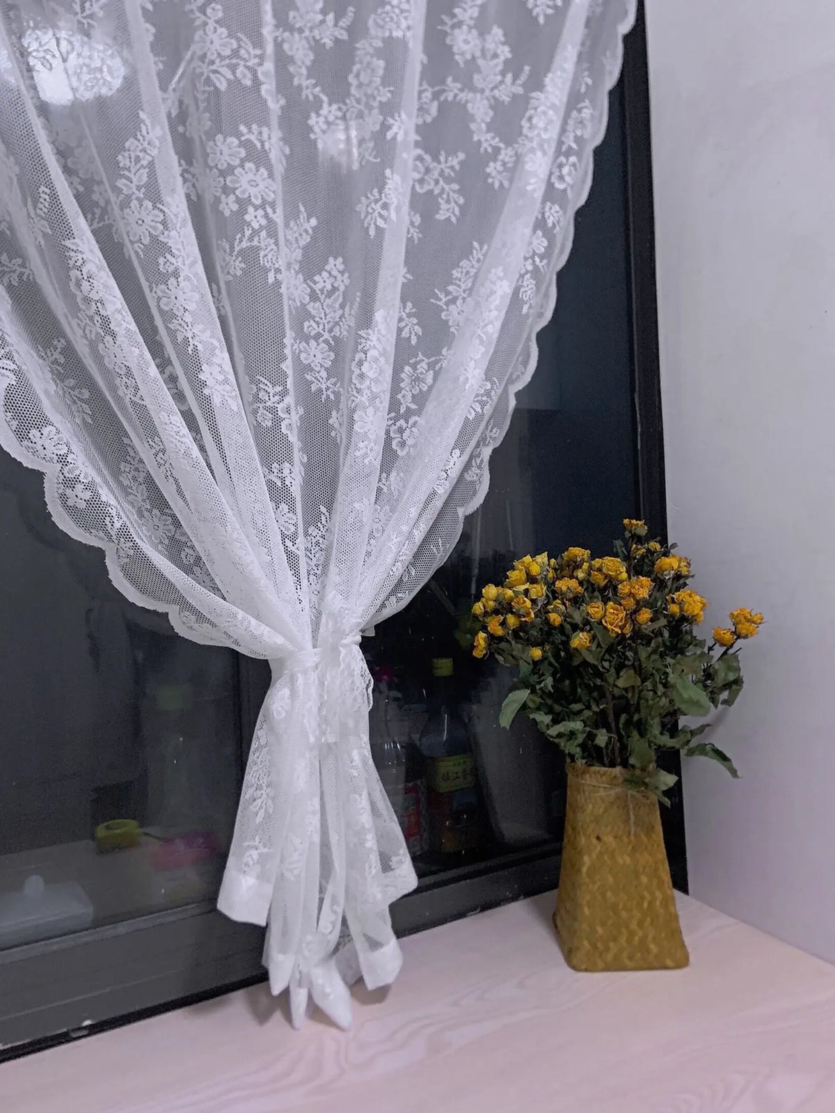 White floral lace sheer curtain panel draped over a glass surface with a vase of yellow flowers.