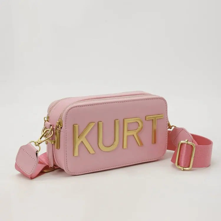 Pinkes Lederbeuteltasche mit goldenen Buchstaben KURT und passende Trageleiste.