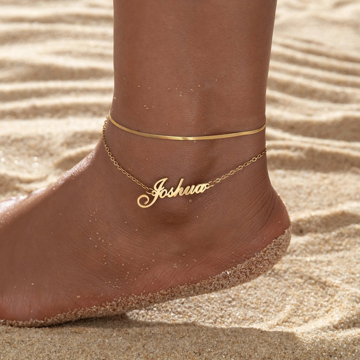 A close-up of a gold double-layer name anklet worn on a person's foot against a sandy background.