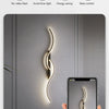 A modern minimalist LED wall lamp illuminates a vertical, wavy light design on a gray wall next to a hand holding a smartphon