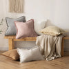 A collection of neutral-toned boho fringe throw pillows and a cream knitted blanket displayed on a wooden bench against a whi