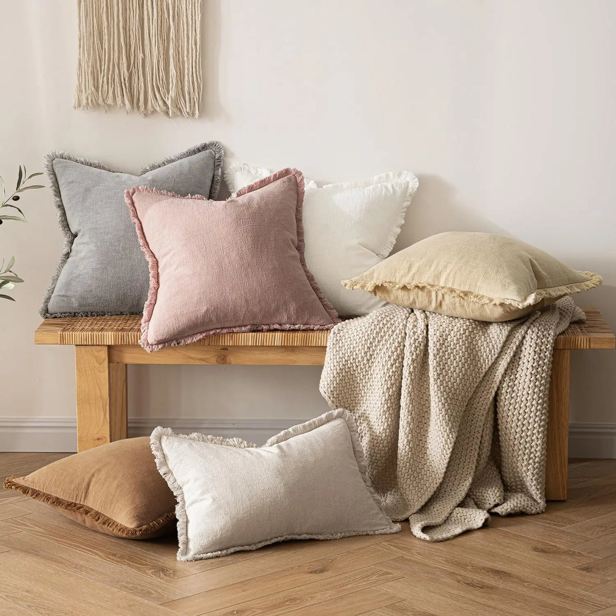 A collection of neutral-toned boho fringe throw pillows and a cream knitted blanket displayed on a wooden bench against a whi