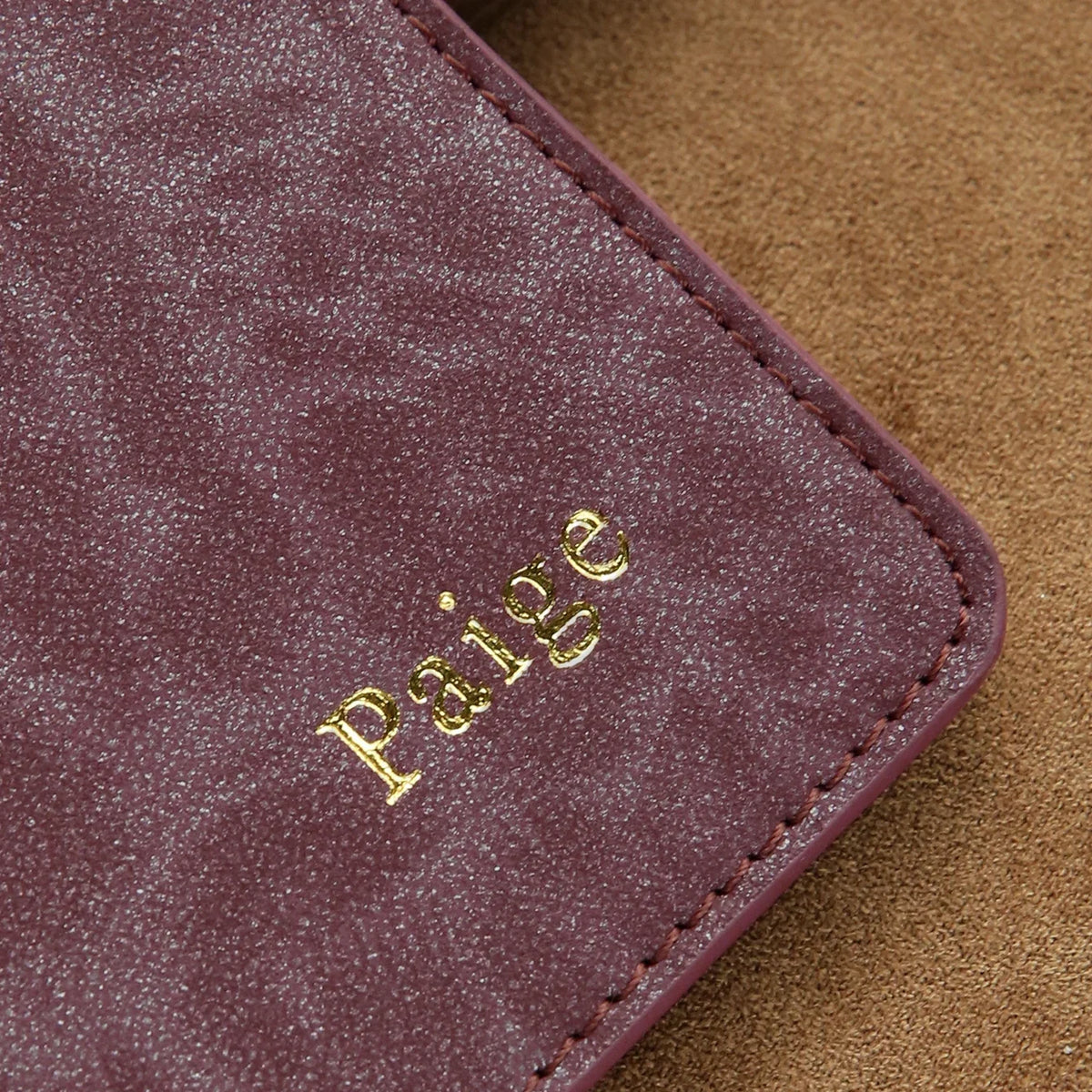 A close-up of a deep purple textured leather passport holder with gold embossed name writing resting on a tan background.