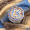 A roll of colorful gradient cake yarn showing various shades of blue, brown, and cream on a fabric background.