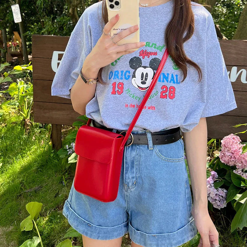 A person is holding a red vintage PU leather crossbody shoulder bag while wearing a light gray t-shirt and denim shorts outdo