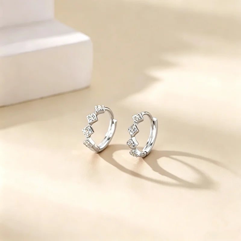 Pendientes de aro plateados con pequeñas piedras de circonita en una superficie beige claro.