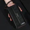 Une main tient un porte-monnaie clutch en cuir noir avec une sangle marron et du texte gravé.