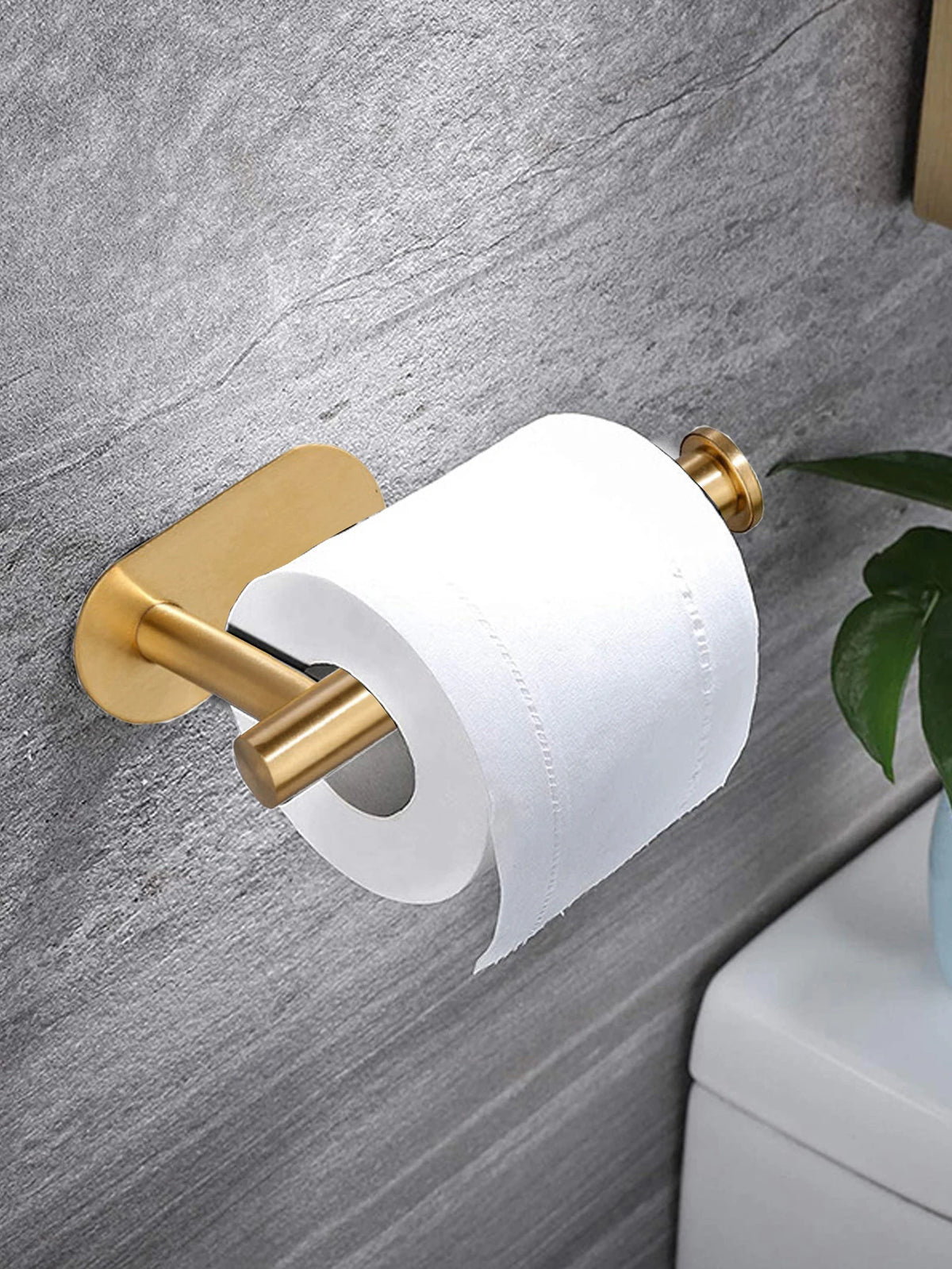 A brass adhesive stainless steel toilet paper holder with a white roll mounted on a gray wall.