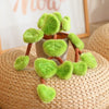 A lifelike plush potted plant with soft green leaves and brown stems sitting on a woven surface.