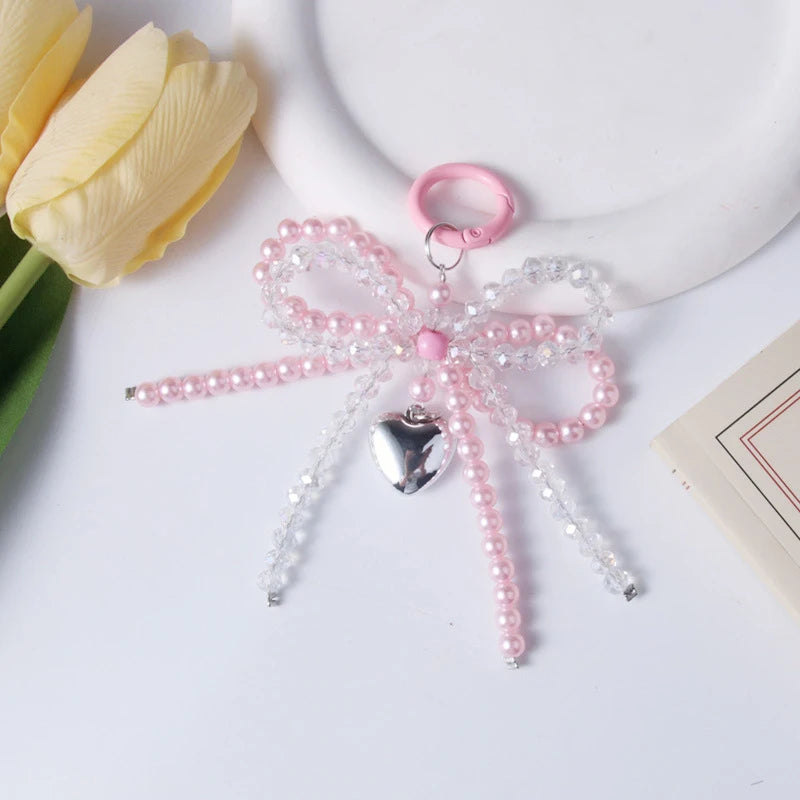 A pink pearl bow keychain with a heart charm is displayed next to a yellow tulip.