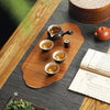 A wooden tea mat holds a ceramic teapot, small cups, and a tea infuser on a dark gray surface.
