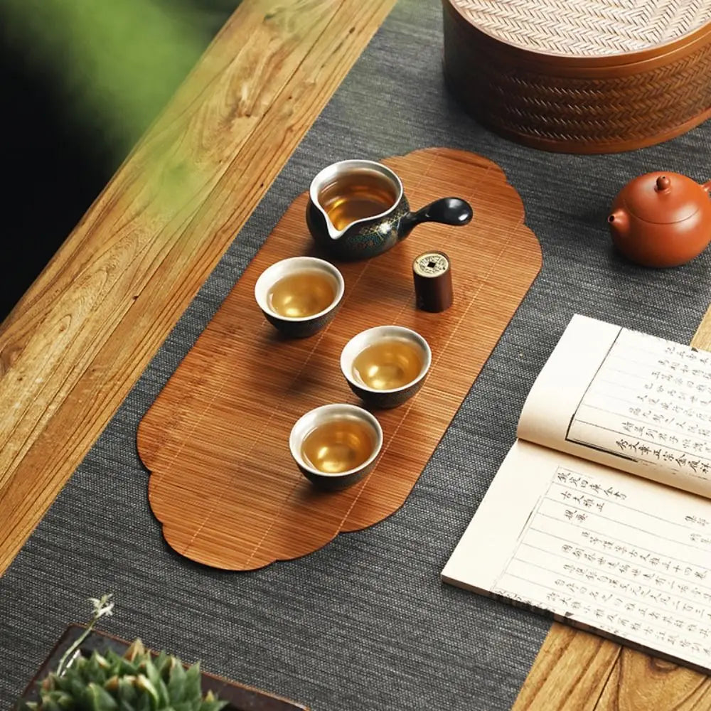 A wooden tea mat holds a ceramic teapot, small cups, and a tea infuser on a dark gray surface.