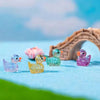 A collection of ten glitter resin duck figurines in various colors are displayed on a blue surface with a wooden archway in t