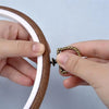 A hand is holding a wooden embroidery hoop with a white inner ring and a brass-colored needle tool.