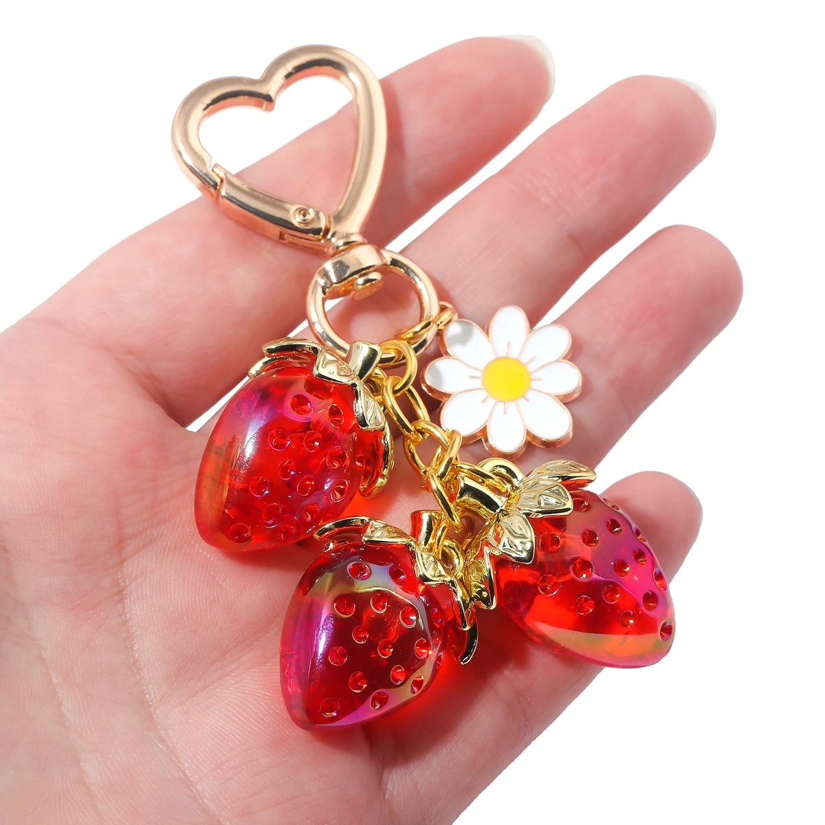 Un charm à clés de fraise de couleur or avec une boucle en forme de cœur et un détail de fleur blanche, tenu dans une main.