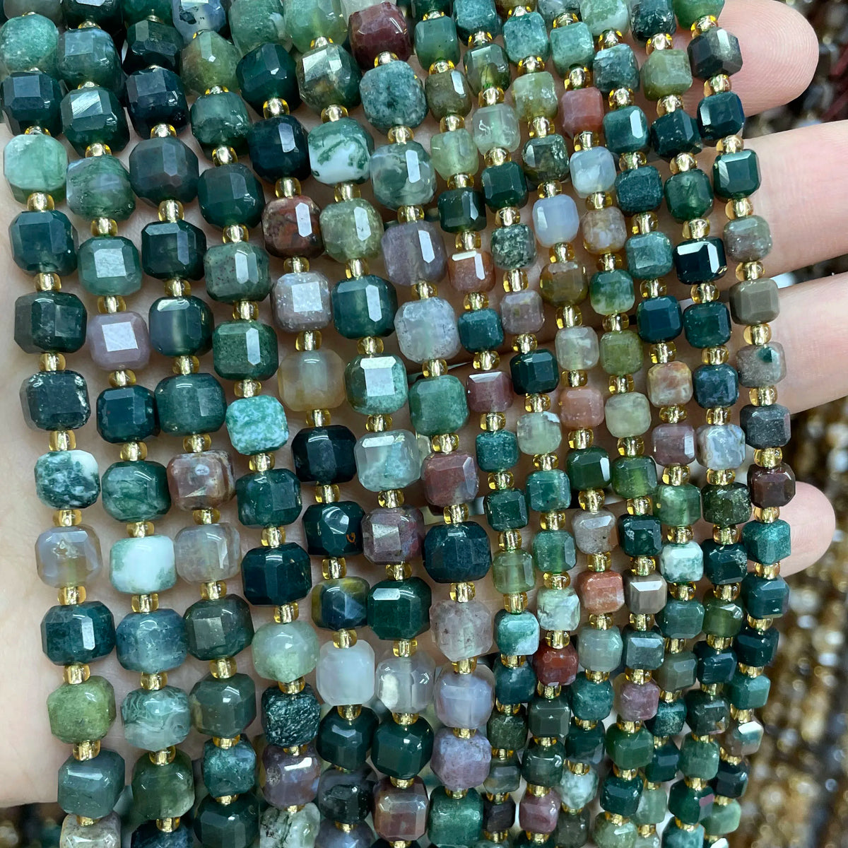 A close-up of natural stone square faceted beads in various shades of green, blue, and pink clustered on a hand.