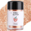 A glass jar of edible glitter powder in rose gold color surrounded by loose rose gold glitter.