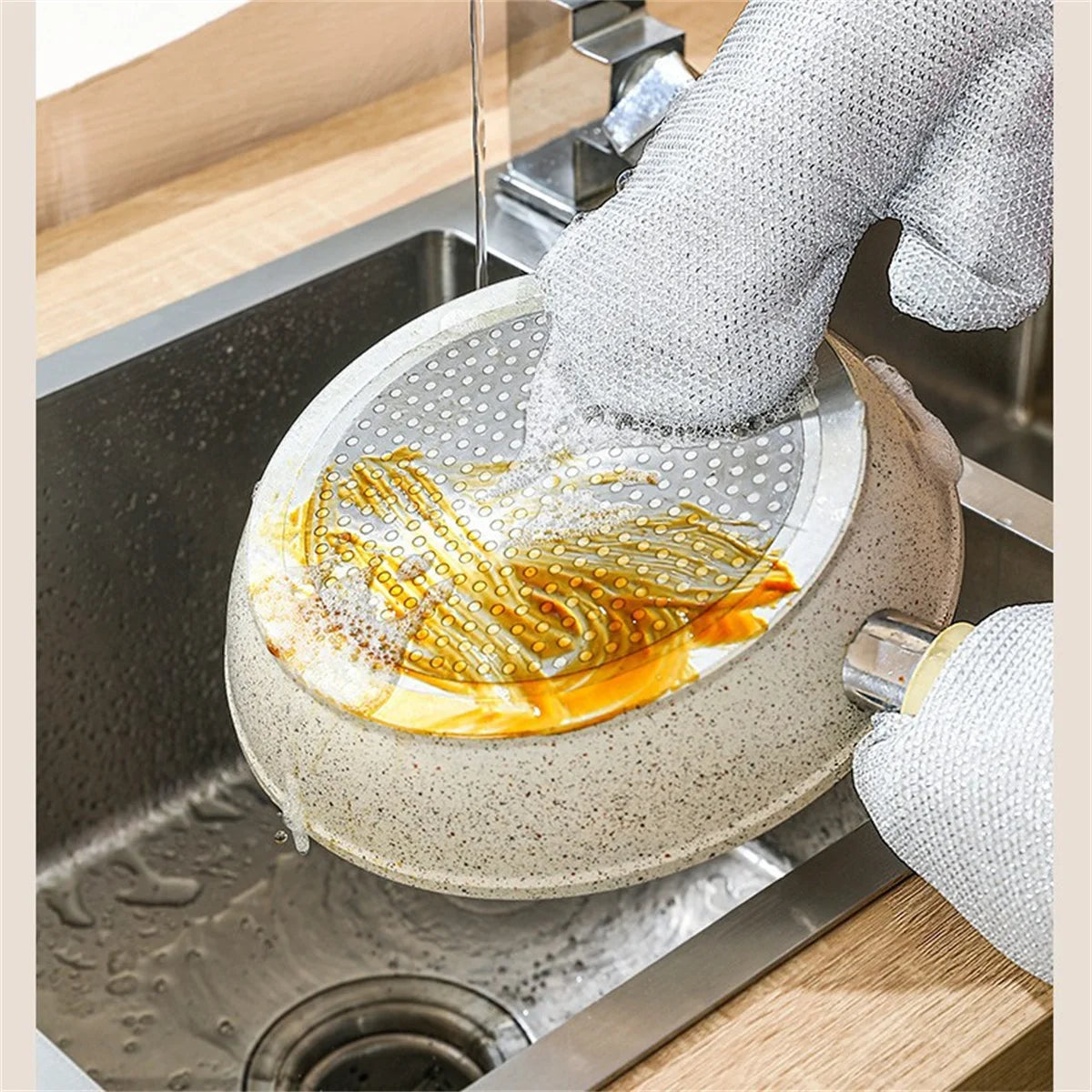 A hand wearing steel-wire dishwashing gloves is washing a plate in a stainless steel sink.