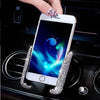 A hand holds a white smartphone with a crystal air vent car phone holder attached, resting on a dark car console.