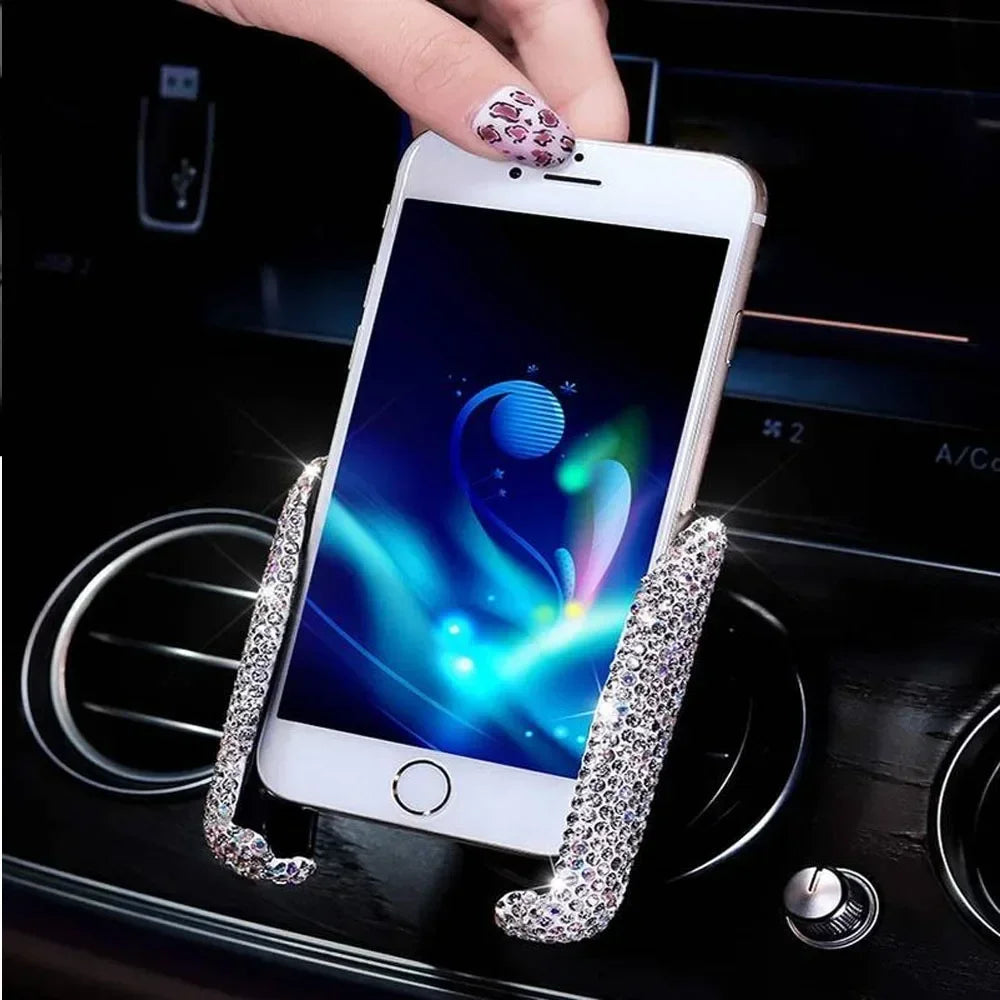 A hand holds a white smartphone with a crystal air vent car phone holder attached, resting on a dark car console.