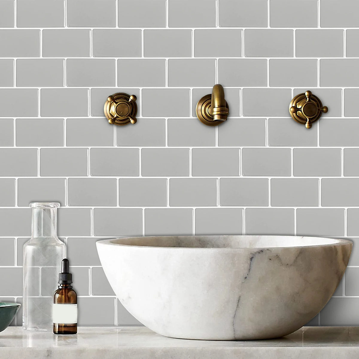 A white marble vessel sink is displayed in front of a gray subway tile backsplash with brass wall hardware.