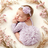 A newborn baby wearing a lavender lace romper and matching headband, surrounded by dried pink floral branches.