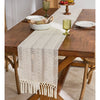 A beige hand-woven table runner with tassels is placed on a dark wood table in a rustic home setting.