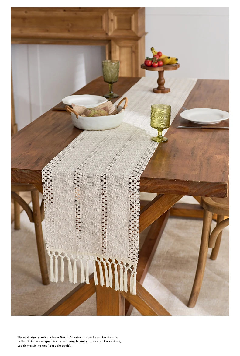 A beige hand-woven table runner with tassels is placed on a dark wood table in a rustic home setting.
