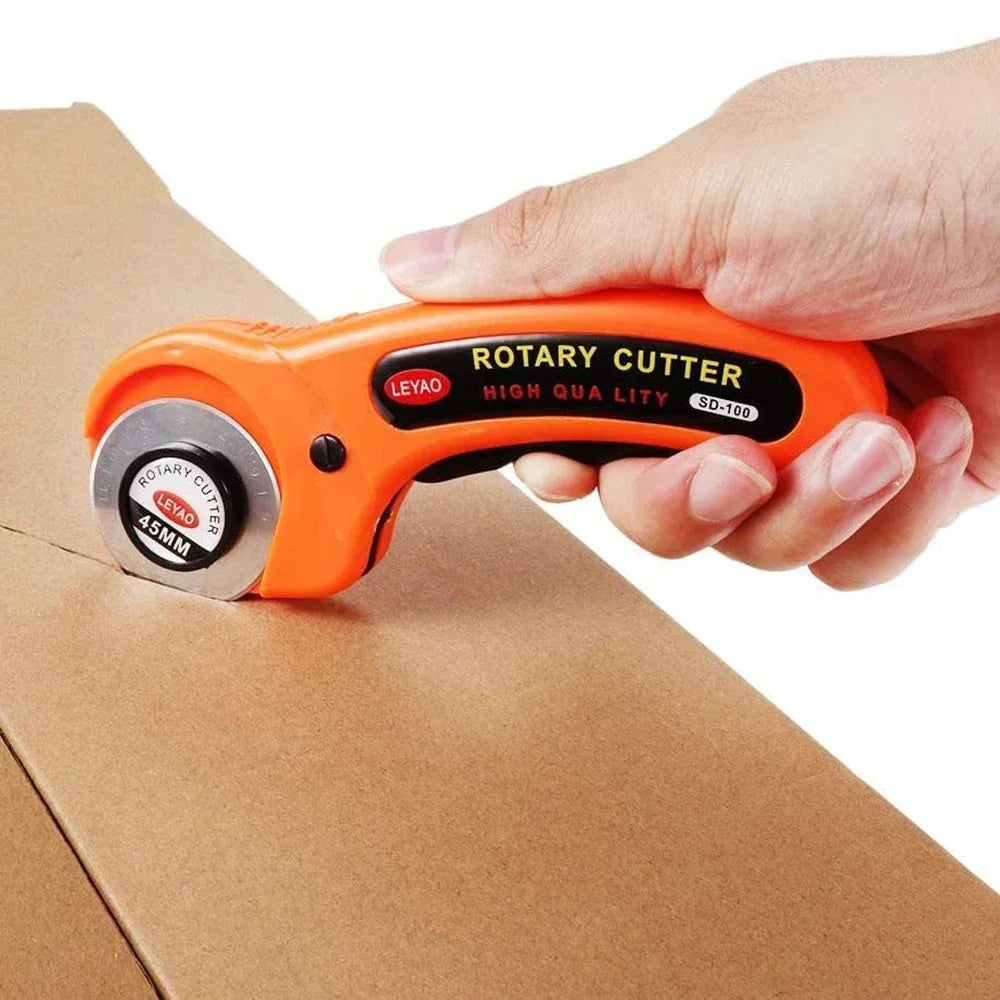 An orange rotary cutter tool is held by a hand, resting on brown cardboard.