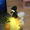 A white bendable robot night light with a glowing head sits on a wooden desk next to a laptop.